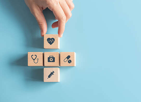 Foto: Eine Hand vor hellblauem Untergrund, die einen Holzbaustein an vier andere anlegt, diese Bausteine haben Symbole aus der Pflege wie ein Herz, eine Spritze, Pillen, ein Stethoskop und ein Arztkoffer