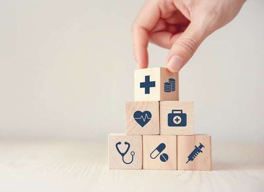 Eine Hand, die eine Pyramide mit Würfel gestapelt hat.. Die Würfel sind mit Pflegesymbole (Spritze, Herz, Tablette ....) bedruckt.